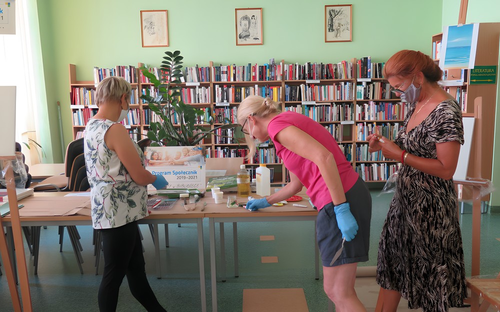 Na tle regałów z książkami i stołu z akcesoriami malarskimi stoją uczestniczki warsztatów. Od lewej: Iwona Sołoducho, Elżbieta Knitter, Elżbieta Żbikowska.