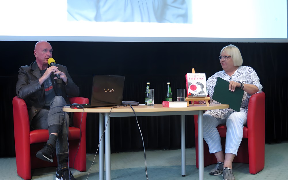 Na zdjęciu od lewej Cezary Harasimowicz siedzi na czerwonym fotelu, dalej prowadząca spotkanie Magda Omilianowicz siedzi również na czerwonym fotelu. Między nimi stoi owalny stolik.