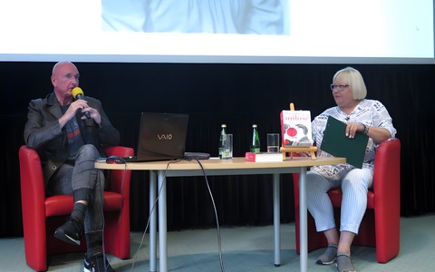 Na zdjęciu od lewej Cezary Harasimowicz siedzi na czerwonym fotelu, dalej prowadząca spotkanie Magda Omilianowicz siedzi również na czerwonym fotelu. Między nimi stoi owalny stolik.