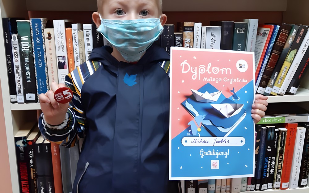 Na zdjęciu Michaś trzymający dyplom w ręku i odznakę akcji - Mała książka Wielki człowiek, w tle książki