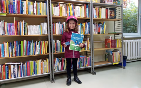 Dziewczynka trzyma w rękach dyplom oraz kartę akcji Mała Książka-Wileki Człowiek. W tle regał z książkami.