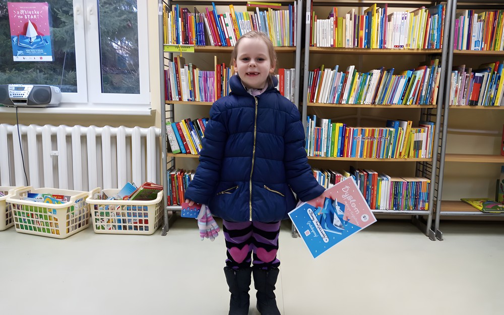 Dziewczynka stoi, trzymając w prawej ręce kartę z naklejkami a w lewej dyplom akcji Mała Książka - Wielki Człowiek. W tle regały z książkami.