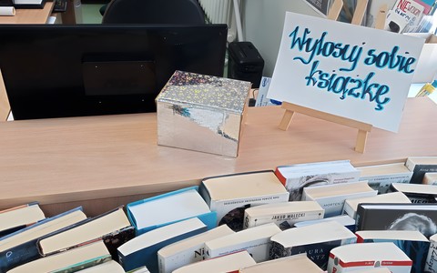 Na biurku stoi błyszczące pudełko obok stojak z białą kartką z czarno-żółtym napisem. Poniżej stoją książki.
