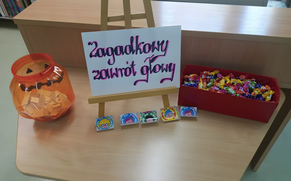 Na stoliku po lewej stronie stoi pomarańczowy pojemnik z białymi karteczkami, w środku znajduje stojak, na którym stoi biała kartka z czarno-różowym napisem: Zagadkowy zawrót głowy, a po prawej stronie widoczny jest czerwony pojemnik wypełniony cukierkami