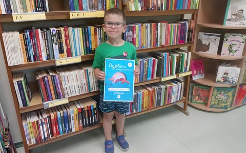 Chłopiec trzymający w rękach dyplom akcji "Mała książka - Wielki Człowiek", w tle regał z książkami