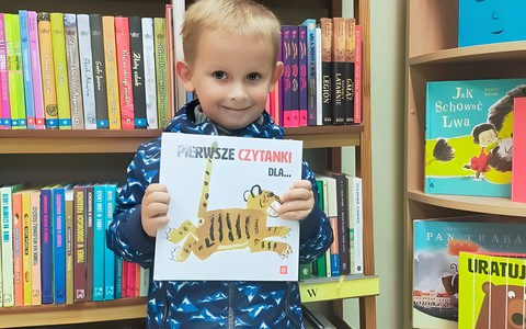 Dziecko trzyma w rękach książeczkę. W tle regał z książkami