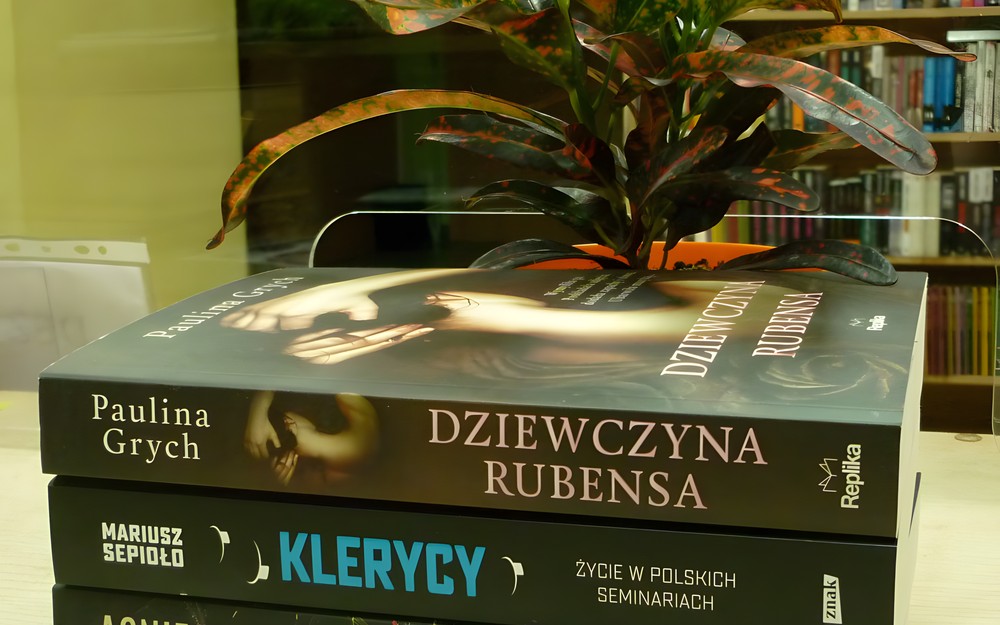 na tle regałów z książkami i kwiatka w doniczce leży stos książek, patrząc od góry: Dziewczyna Rubensa autorstwa Grych P., Klerycy autorstwa Sepilo M., Kto jest bez grzechu autorstwa Jeż A. i Kochana córeczka autorstwa Ragsdale A.