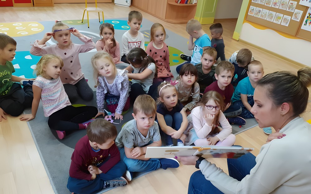 Bibliotekarka czyta dzieciom książkę. Na drugim planie grupa przedszkolaków siedząca na dywanie