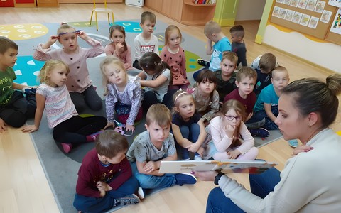 Bibliotekarka czyta dzieciom książkę. Na drugim planie grupa przedszkolaków siedząca na dywanie