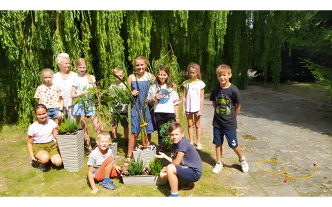 Bibliotekarka Urszula w otoczeniu dzieci, podczas zajęć przyrodniczych w ramach wakacji w bibliotece w ogrodzie filii nr 9 KBP