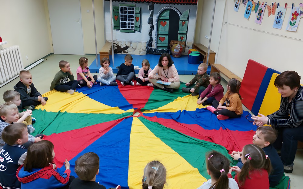 Dzieci podczas zajęć siedzą w kole na kolorowej chuście i słuchają opowiadania, czytanego przez bibliotekarkę