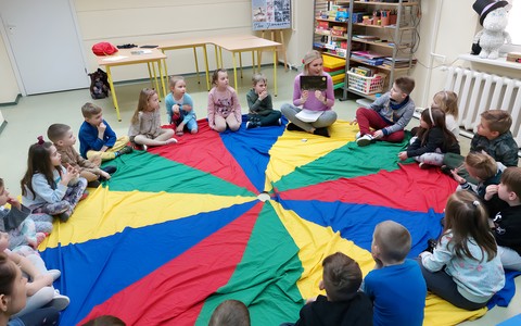 Dzieci siedzą na chuście wraz z bibliotekarką, która prezentuje książkę