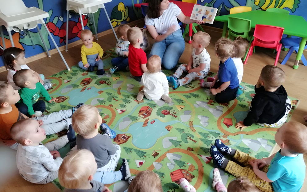 Bibliotekarka czyta dzieciom książkę, dzieci siedzą na kolorowym dywanie