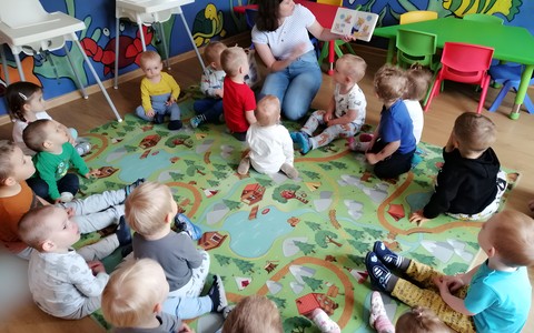 Bibliotekarka czyta dzieciom książkę, dzieci siedzą na kolorowym dywanie