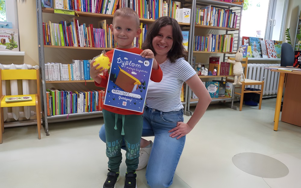 Chłopiec stoi i trzyma dyplom, obok kuca bibliotekarka; w tle książki