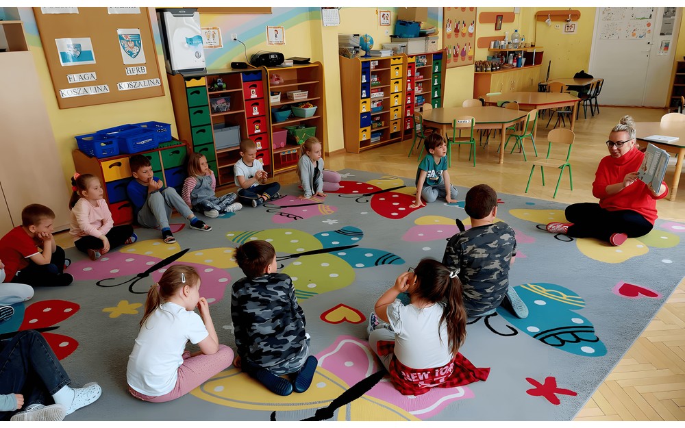 Dzieci siedzą na podłodze i słuchają czytającej książeczkę pt. "Poszukiwany tajemniczy złodziej książek" Gosi