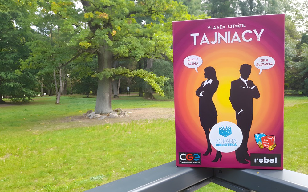 Opakowanie gry Tajniacy na balustradzie, w tle park.