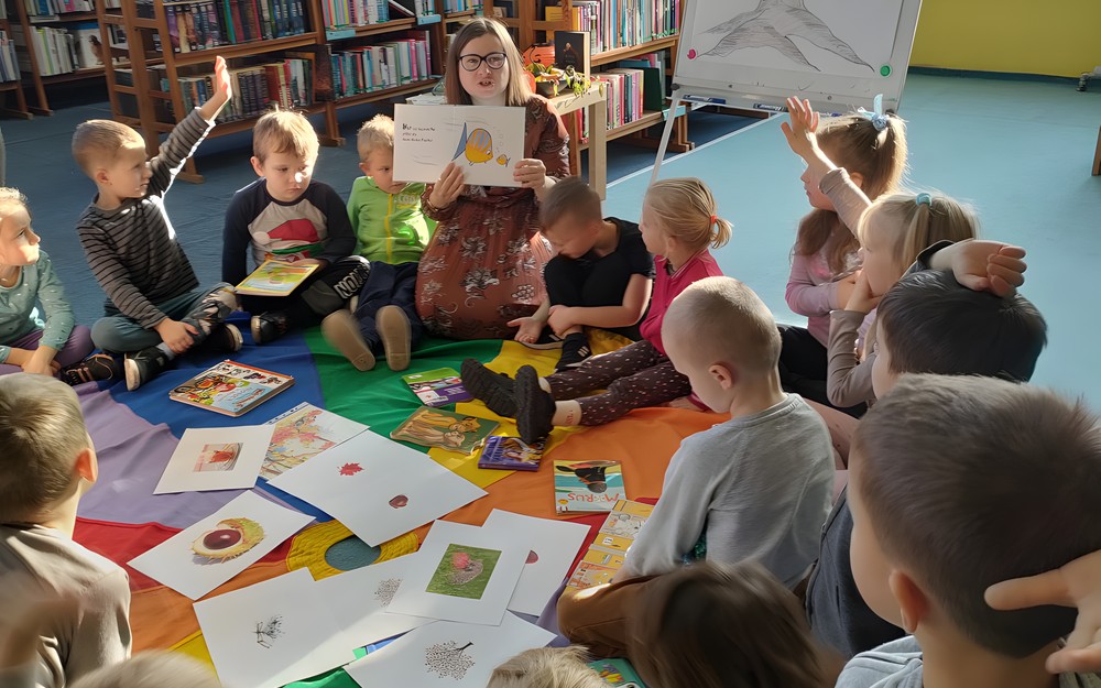 Dzieci siedzą na chuście animacyjnej i słuchają czytanego przez bibliotekarkę opowiadania