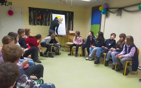 Uczniowie siedzą na krzesłąch słuchając bibliotekarki