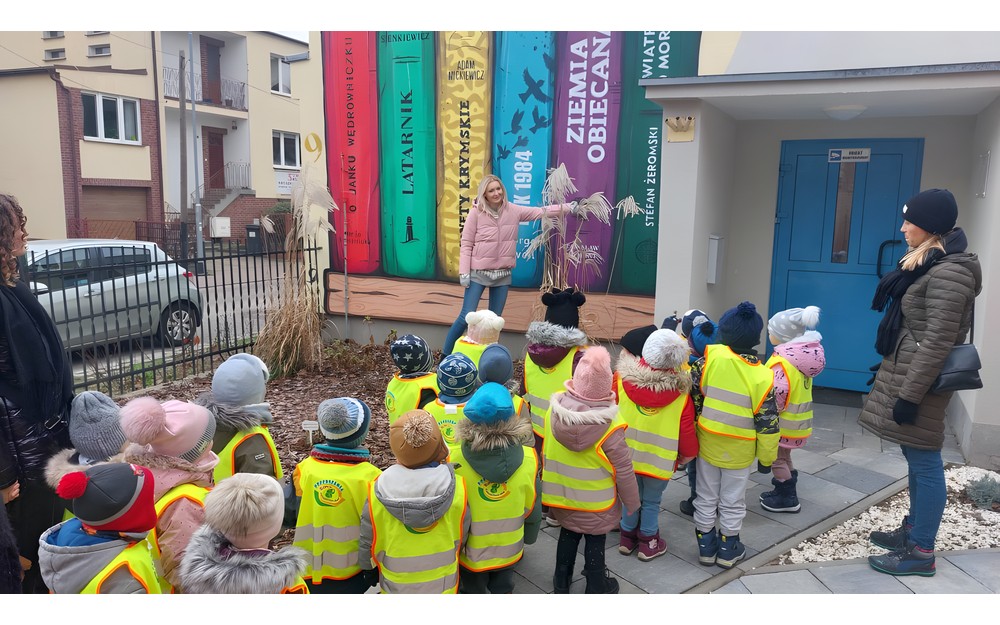 Dzieci stoją przd biblioteką słuchając bibliotekarki