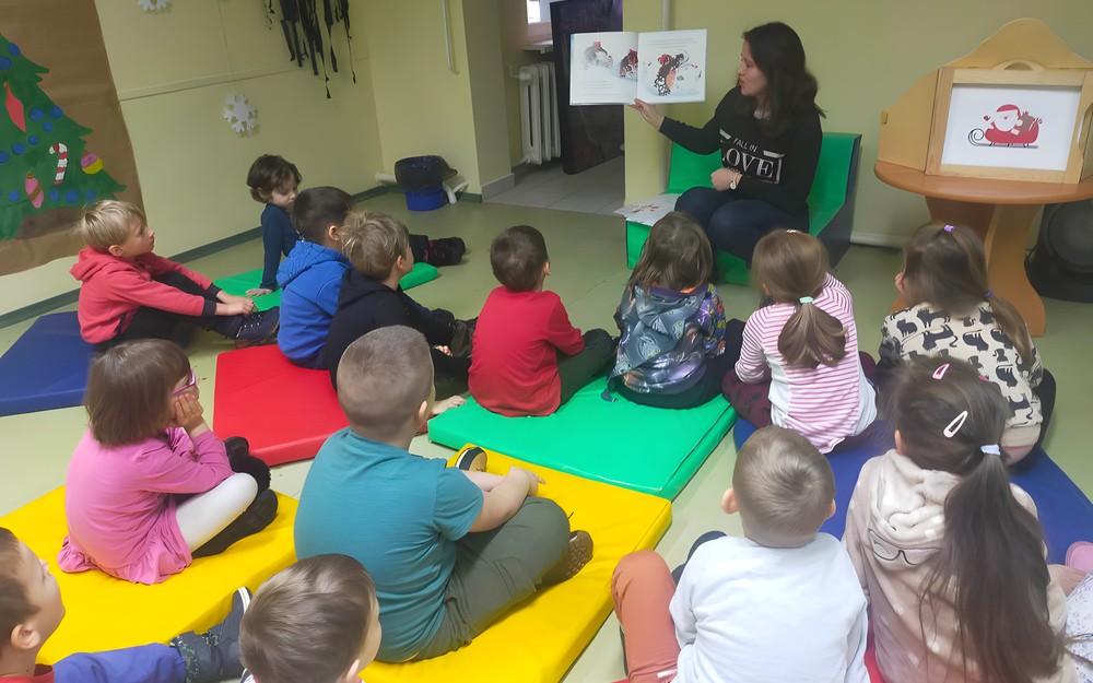 Dzieci podczas zajęć siedzą w kole na kolorowej chuście i słuchają opowiadania, czytanego przez bibliotekarkę