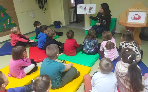 Dzieci podczas zajęć siedzą w kole na kolorowej chuście i słuchają opowiadania, czytanego przez bibliotekarkę