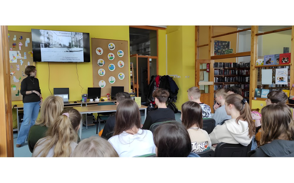 mlodzież siedzi na krzesłach, podczas gdy Pani Soraya Kuklińska opowiada o Powstaniu Warszawkim