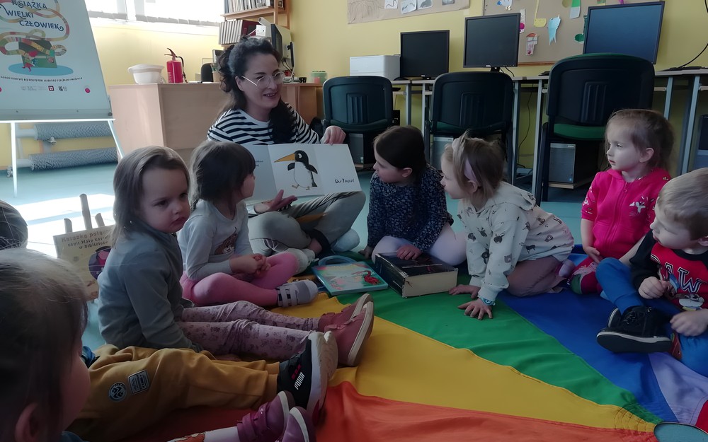 bibliotekarka pokazuje dzieciom siedzącym na chuście amimacyjnej