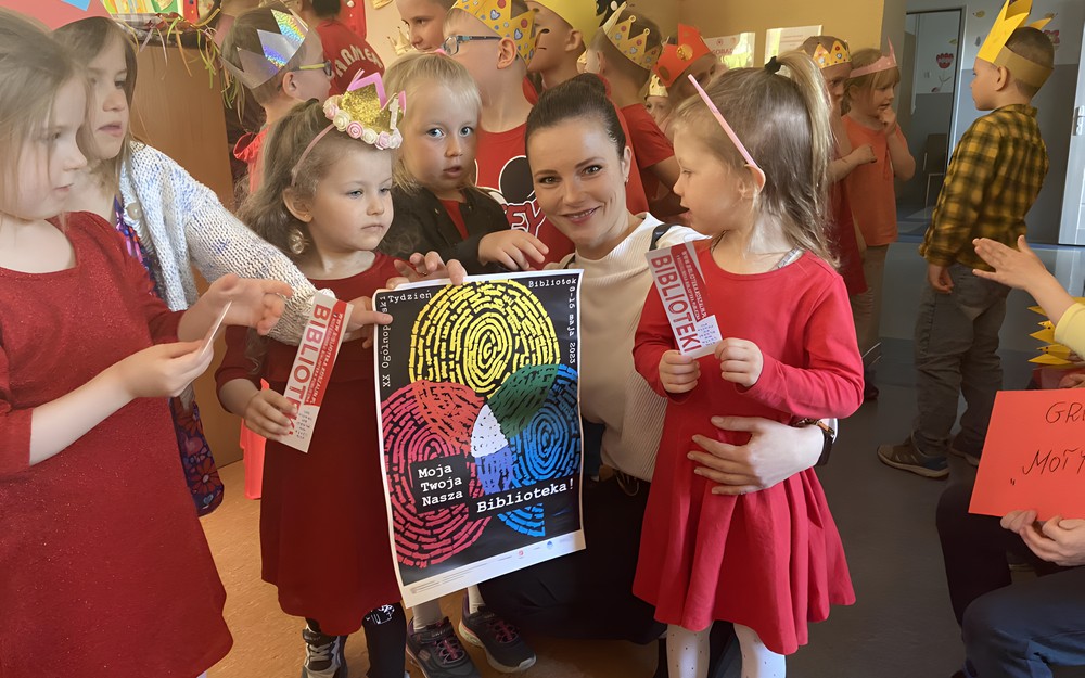 Bibliotekarka z dziećmi prezentuje plakat promujący Tydzień Bibliotek