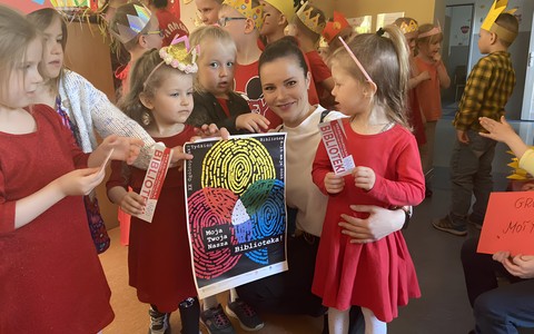 Bibliotekarka z dziećmi prezentuje plakat promujący Tydzień Bibliotek