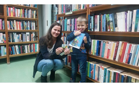 Bibliotekarka z chłopcem prezentują książkę, w tle regały z książkami