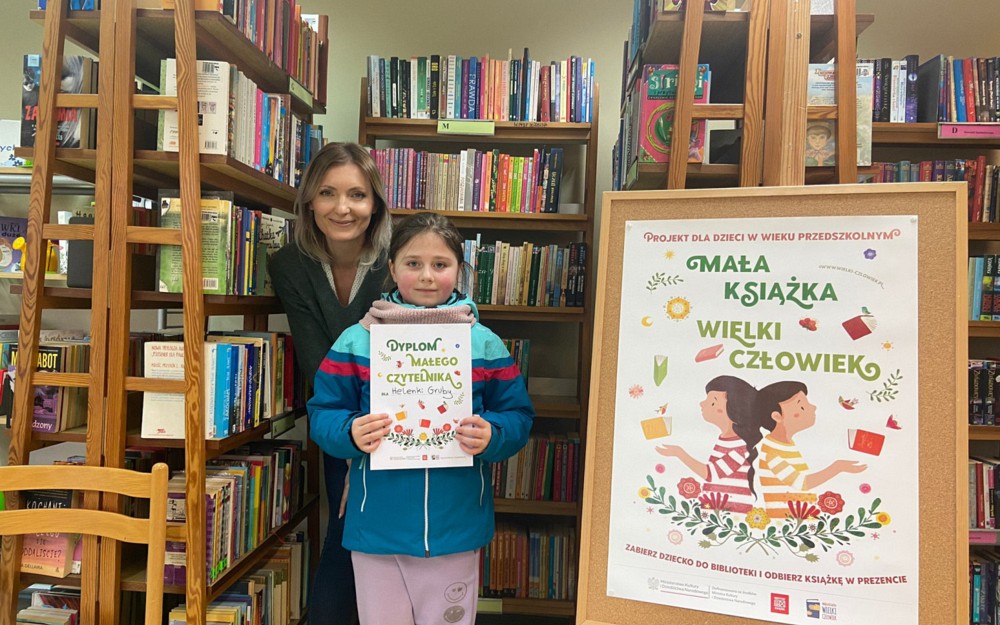 Dziewczynka pozuje z dyplomem, za nią stoi bibliotekarka