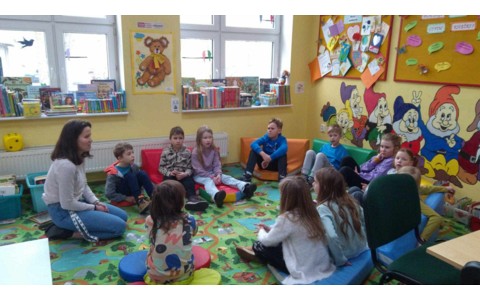 Dzieci siedzą na kolorowym dywanie i słuchają bibliotekarki opowiadającej historie