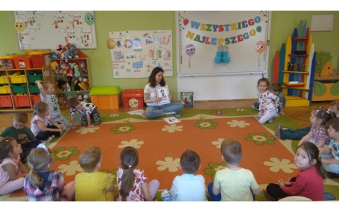 Dzieci siedzą na kolorowym dywanie a bibliotekarka pokazuje dzieciom obrazek