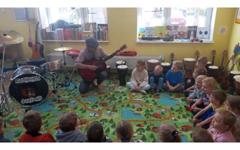 Pan Wojtek gra na gitarze, a dzieci siedzą na dywanie i słuchają kojącej muzki