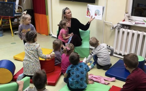Dzieci siedzą na kolorowych pufach i słuchają bajki czytanej przez bibliotekarkę
