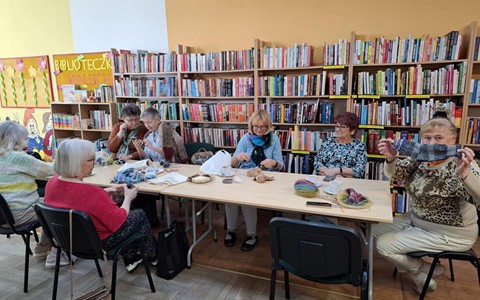 Panie siedzą przy stole, robią na drutach, rozmawiają. Jedna z Pań pokazuje skarpetkę zrobioną na drutach