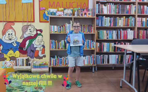 Chłopiec trzyma niebieską książkę;  w tle regał z książkami.