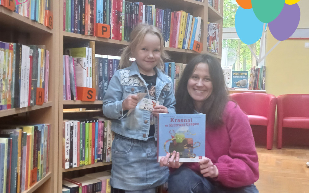 Bibliotekarka wspólnie z dziewczynką trzyma książkę i kartę czytelnika