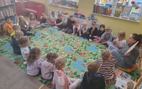 Bibliotekarka wspólnie z dziećmi siedzi na dywanie i czyta opowiadanie