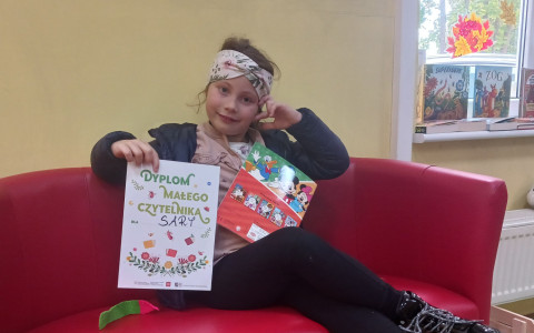 Dzieczynka siedzi na kanapie i prezentuje dyplom super czytelnika
