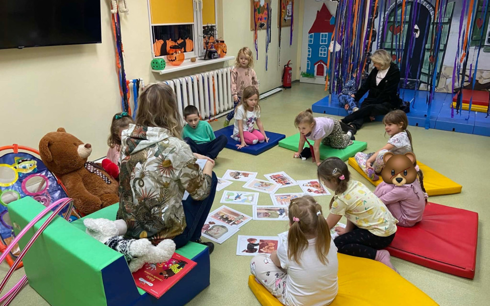 Dzieci siędzą z biblioterką w kółku i słuchają historii powstania pluszowego misia