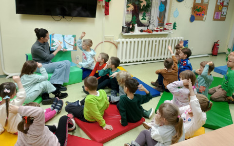 Dzieci siedzą przed panią bibliotekarką na materacykach. Pani bibliotekarka czyta książkę, prezentując jej obrazki dzieciom. Dzieci mają rączki w górze.