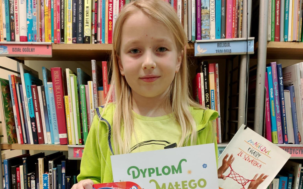 Helenka trzyma uzyskany Dyplom Małego Czytelnika oraz mini prezent od Pań bibliotekarek- mini kręgle. W tle widać regał z książkami.