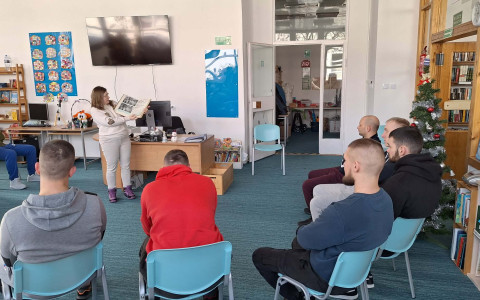 Bibliotekarka opowiada osadzonym o historii biblioteki, trzymając w ręku kronikę z ciekawostkami