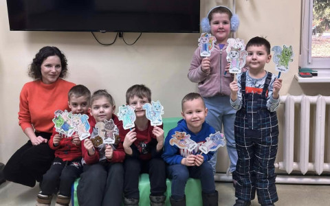 Bibliotekarka i dzieci iedzą na materacyku i pozują do zdjęcia. Dzieci trzymają własnoręcznie zrobione zakładki.