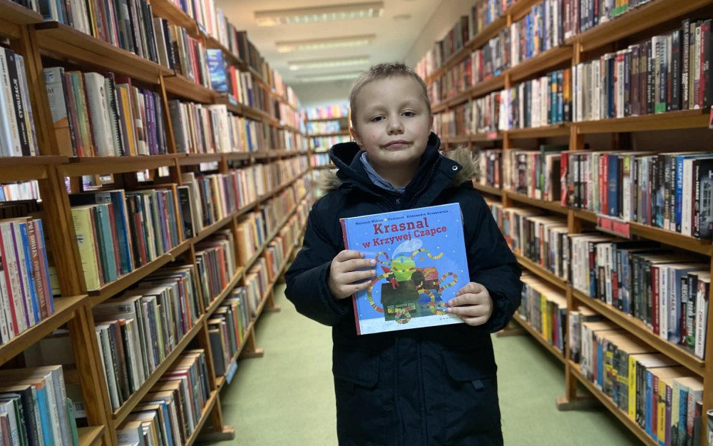 Chłopiec trzyma w rękach książkę i pozuje do zdjęcia na tle regałów z książkami