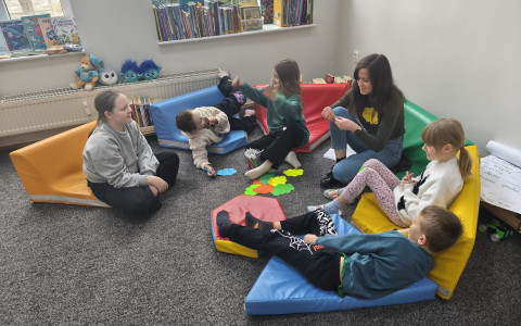 Piątka dzieci wraz z bibliotekarką siedzą na kolorowych matarcach i rozwiązuja zagadki o superbohaterach