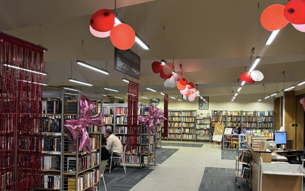 Wnętrze biblioteki – przestronna sala z regałami pełnymi książek ustawionymi w równych rzędach. Przy suficie zawieszone są czerwone, różowe i białe balony oraz dekoracyjne, czerwone, połyskujące kurtyny. Na regałach umieszczono różowe balony w kształcie gwiazd. W głębi sali znajduje się stanowisko bibliotekarza z biurkiem i komputerem oraz stoliki dla czytelników. Oświetlenie stanowią podłużne lampy sufitowe. Podłoga jest jasna, z ciemniejszymi wykładzinami między regałami.