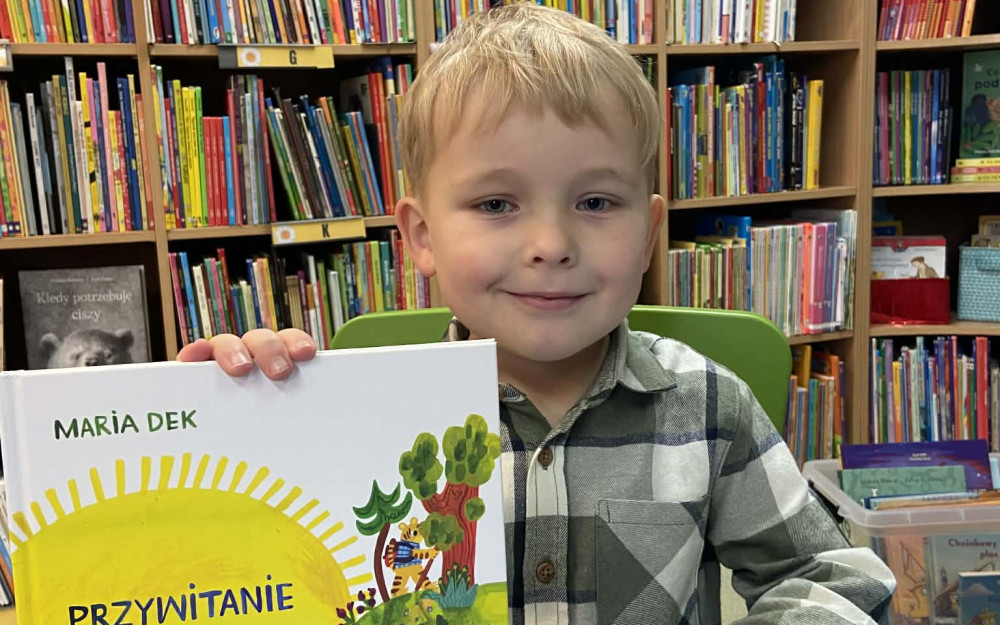 Chłopiec uśmiecha sie do aparatu. w ręku trzyma książeczkę.